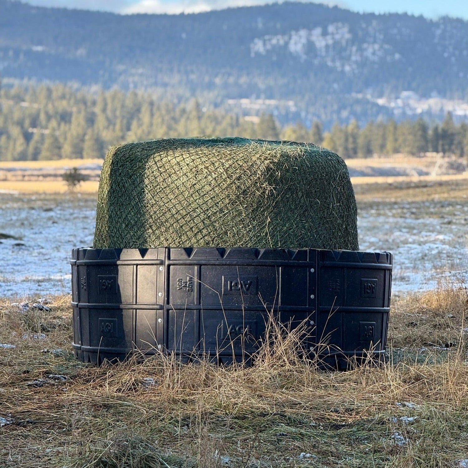 Black plastic haybale feeder with netting on top in a field with mountains in the background