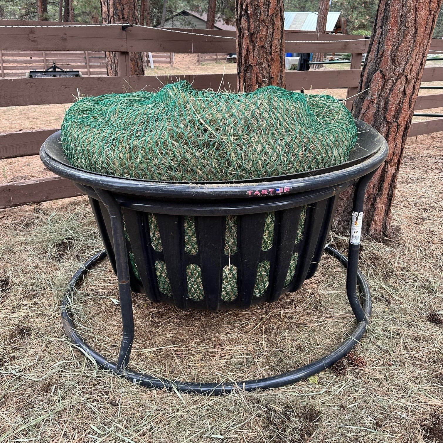 A black tarter basket hay net filled with green hay, placed on a grassy area next to a wooden fence in a pasture.